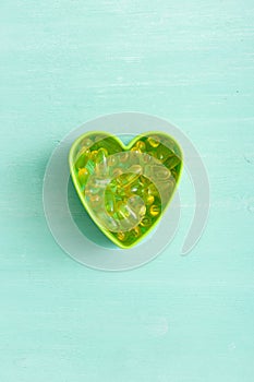 Top view on green plastic heart shape with pile of capsules Omega 3 on turquoise background. fish fat oil capsules