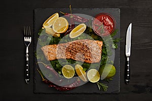 top view of grilled salmon steak, pieces of lime and lemon, chili peppers, sauce and cutlery on black surface