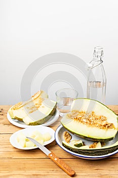 Top view of half green melon, plates with slices and knife on wooden table with bottle and glass of water