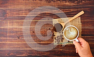 Top view hand holding hot latte coffee in white ceramic cup  with roasted coffee beans  ground coffee on wood spoon on brown sack