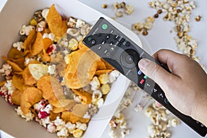 Top view of a hand holding tv remote control against fried popcorn and chips on a bowl
