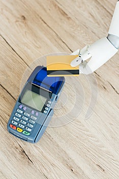 top view of hand of robot holding credit card above payment terminal on wooden