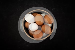 Top view of handmade ceramic bowl of fresh organic brown and white eggs on dark moody background.