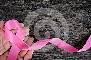 Top view of hands holding bright pink ribbon on dark wood background. Breast cancer awareness and women`s health concept.