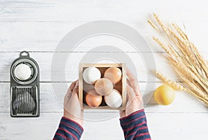 Top view hands holding chicken egg in box, white wood background