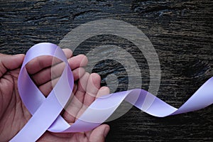 Top view of hands holding lavender or light purple orchid color ribbon on dark wooden background. General and testicular cancer