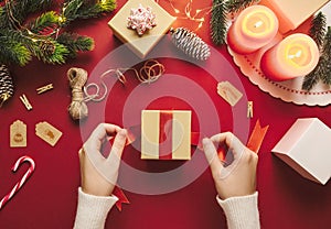 Top view of hands preparing and wrapping Christmas gift box. Merry Xmas rustic decor stuff on a red background and winter holiday