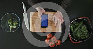 Top view. Hands with the telephone above the cutting board. Vegetables and knife placed nearby.