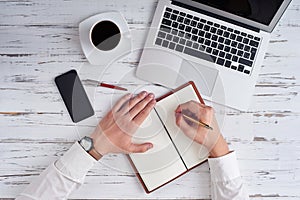 Top view of hands writing in notepad, laptop, phone, pen and glasses on table in office