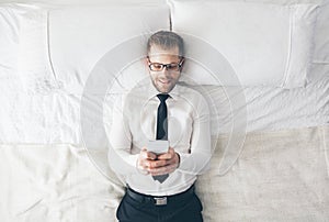 Top view. Handsome businessman with glasses lying on bed texting from his smartphone