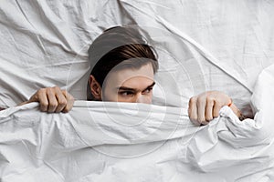 Top view of handsome young man hiding under blanket in bed at home and