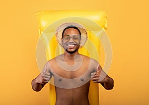 Top view of happy black man lying on inflatable lilo and showing thumbs up with both hands, yellow background