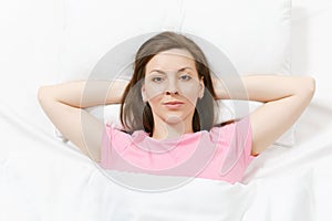 Top view of happy brunette young woman lying in bed with white sheet, pillow, blanket, hands under head. Smiling female