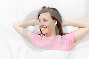 Top view of happy brunette young woman lying in bed with white sheet, pillow, blanket, hands under head. Smiling female
