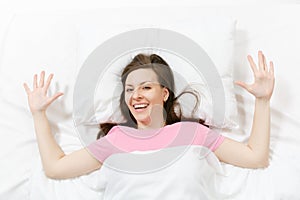 Top view of happy brunette young woman lying in bed with white sheet, pillow, blanket. Smiling female spreading hands