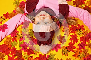 Top view of a happy little girl lying on autumn leaves. The child is upside down in a scarf and hat