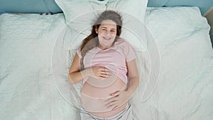 Top view of happy smiling pregnant woman enjoying pregnancy lying on bed and stroking her big belly. Concept of