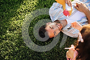 Top view of happy young father playing with his daughter with bird toy lying on the green grass outdoors. Dad enjoying at day out