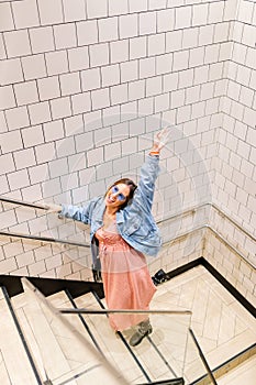 Top view of happy young woman on urban clothes climbing stairs. White tile wall. Fashion model wearing pink dress, denim jacket,
