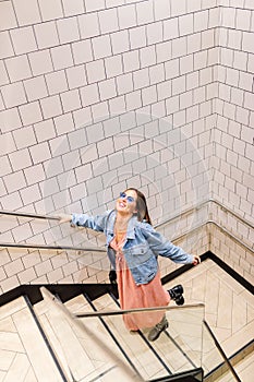 Top view of happy young woman on urban clothes climbing stairs. White tile wall. Fashion model wearing pink dress, denim jacket,