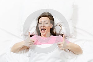 Top view of head of happy brunette young woman lying in bed with white sheet, pillow, blanket. Smiling pretty female