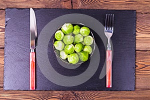 Top view on healthy fresh Brussels sprouts, knife and fork on a black slate on a wooden table - vegan concept