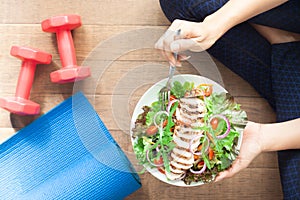 Top view healthy girl eating chicken salad. Dumbbells and yoga mat on the floor