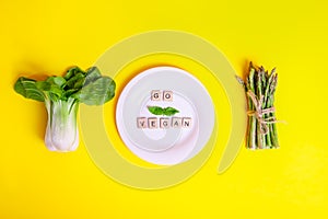 Top view healthy Pak Choi, asparagus and white plate with Go vegan message on wooden blocks on the yellow background. Vegetarian