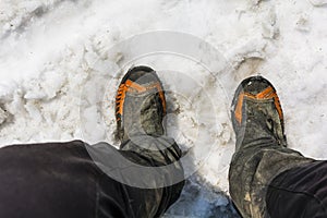 Top view of hiking boots on the feet of a tourist and soiled pants during a mountain trip in the spring snow