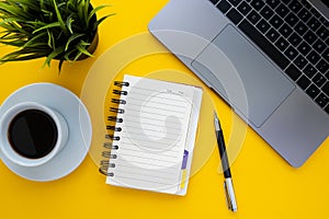 Top view of home office desk. Laptop, empty notebook, pen, cup of coffee, green plant on yellow background.