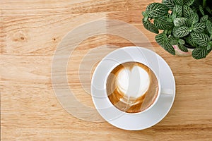 Top view of hot coffee latte with heart shaped milk foam on light wood table background with tabletop small pot of Fittonia nerve