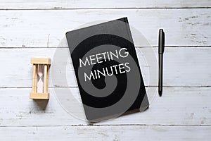 Top view of hour glass,pen and notebook written with 'MEETING MINUTES' on white wooden background