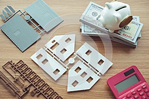 Top view of house model parts, piggy bank, calculator and bill banknotes on wooden table background.