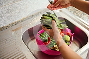 Top view of housewife hand holding artichoke and rinsing it under flowing water in stainless sink in the kitchen at home