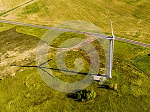 Top view of huge bright green field and lonely white wind turbine that casts shadow parallel to road