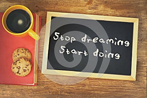 Top view image of blackboard with the phrase stop dreaming start doing, next to coffee cup and cookies