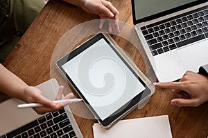 Top view image of a blank screen digital tablet mockup on a meeting table with laptops and papers