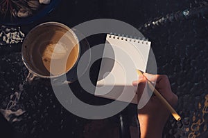 Top view image of a hand holding a pen and writing on a blank white notebook with coffee cup