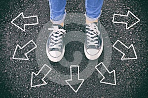 top view image of person in jeans and retro shoes standing over asphalt road with painted arrows showing different directions.