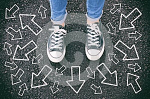 top view image of person in jeans and retro shoes standing over asphalt road with painted arrows showing different directions.