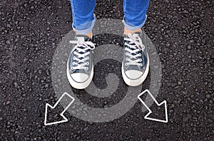 top view image of person in jeans and retro shoes standing over asphalt road with painted arrows showing different directions.