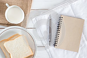 Top view image of Sliced bread on dish with hot milk, pencil and Brown paper notebook on white wood table background, Breakfast in