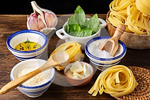 Top view of ingredients for pesto sauce, parmesan cheese, salt, olive oil, basil and garlic with tagliatelle in bowl, wooden spoon