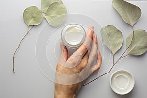 Top view of a jar of cream on a white background, with eucalyptus leaves and womens hand.