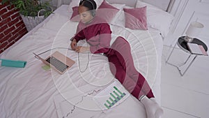 Top view of joyful slim African American young woman lying on bed conferencing online on laptop. Positive confident