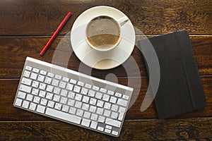 Top view of keyboard, pencil, black notebook and a cup of coffee on a wooden table.