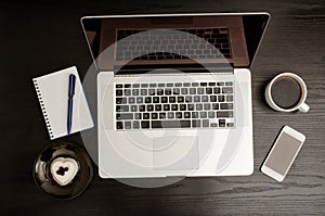 Top view of a laptop keyboard, coffee mug, a smartphone, a notebook and a dessert on a black wooden table