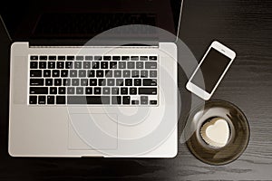 Top view of a laptop keyboard, smartphone and dessert on a black wooden table