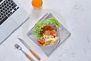 Top view of laptop, orange juice in glass and fresh salad in lunch box at workplace