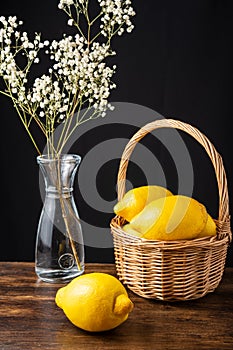 Top view of lemons in basket with handle, bottle with white flowers and whole lemon on wooden table and black background,